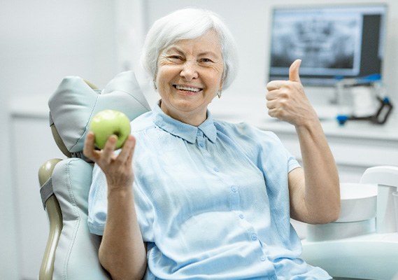 An older, smiling woman holding a green apple and giving a thumbs up