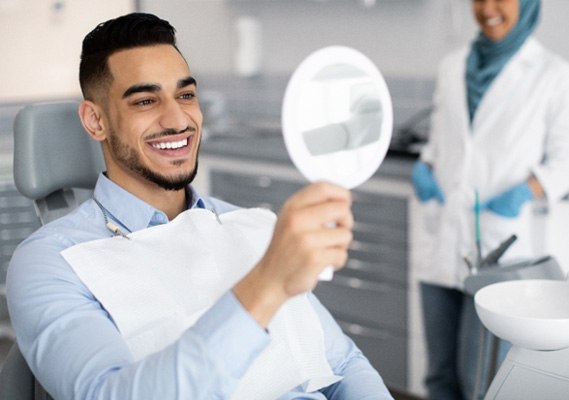 a smiling man looking in a handheld mirror while sitting in a dental chair